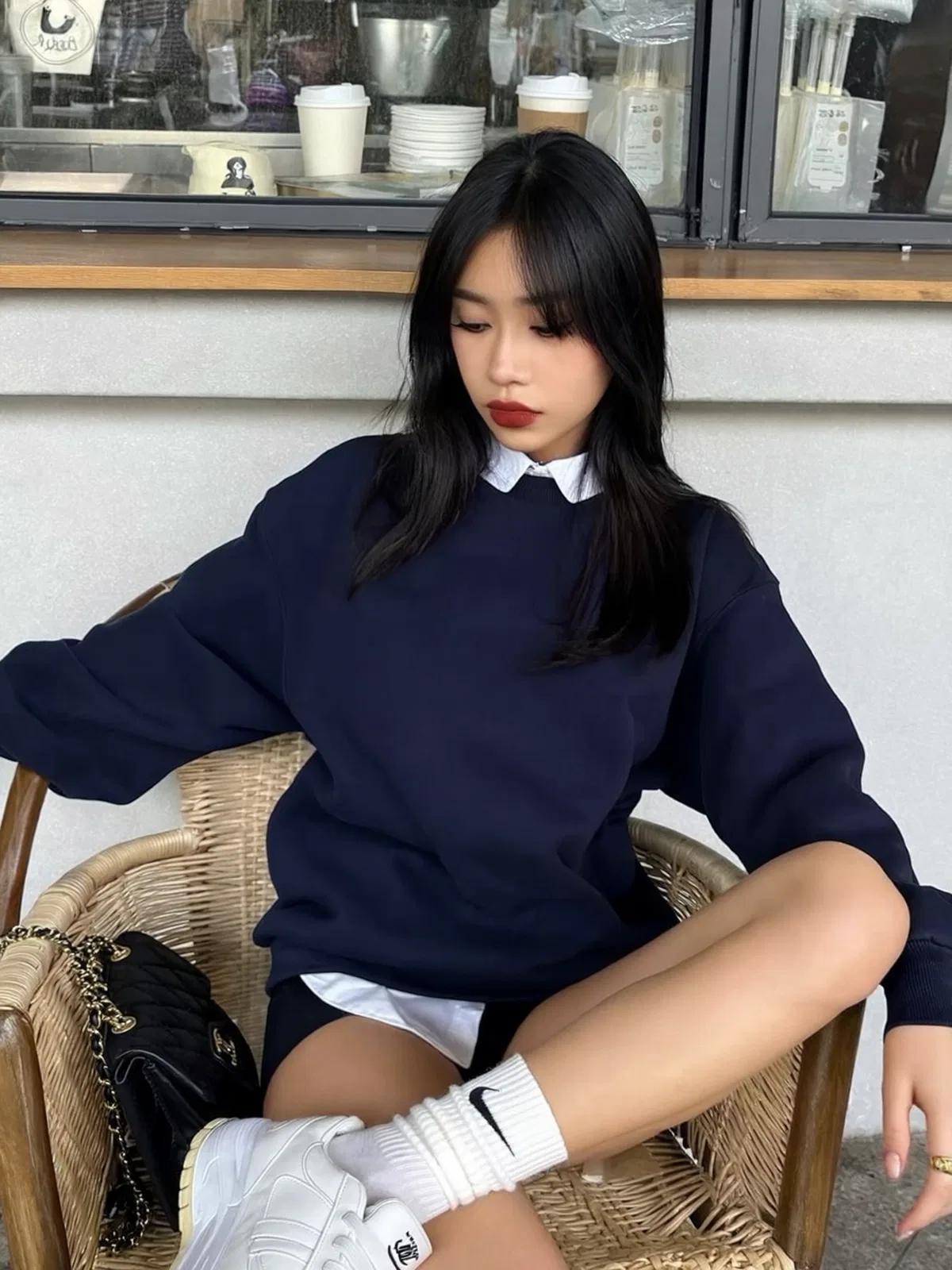 Girl seated in a woven chair at a café, wearing a navy sweater, white collar, white shorts, socks, and sneakers.