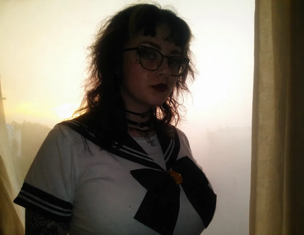Girl with dark wavy hair and glasses wearing a sailor-style top by a bright window