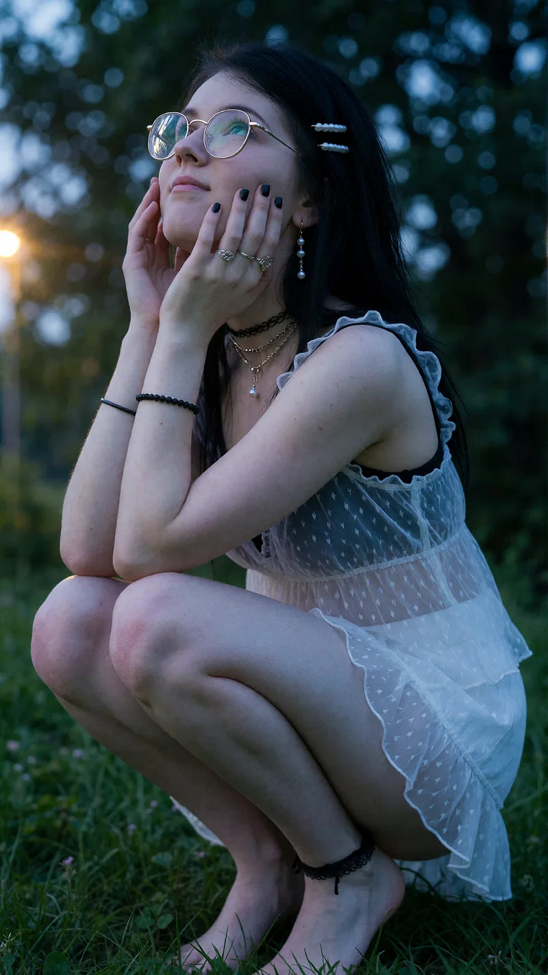 Girl with long dark hair and round glasses crouching on grass in a sheer white ruffled dress at sunset