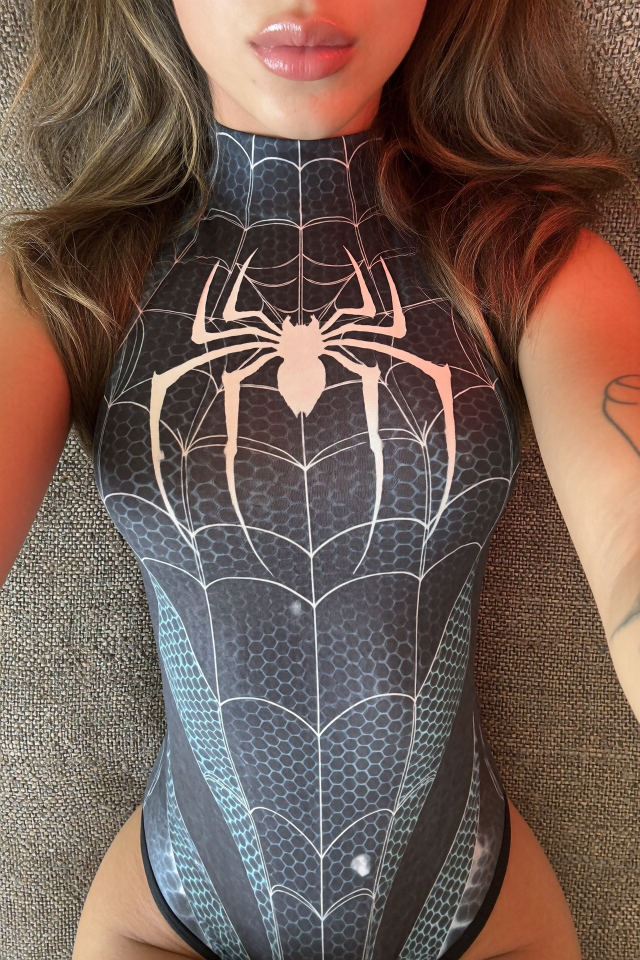 Cropped close-up of a person with wavy brown hair, glossy lips, and a black spiderweb-patterned bodysuit against gray textured fabric