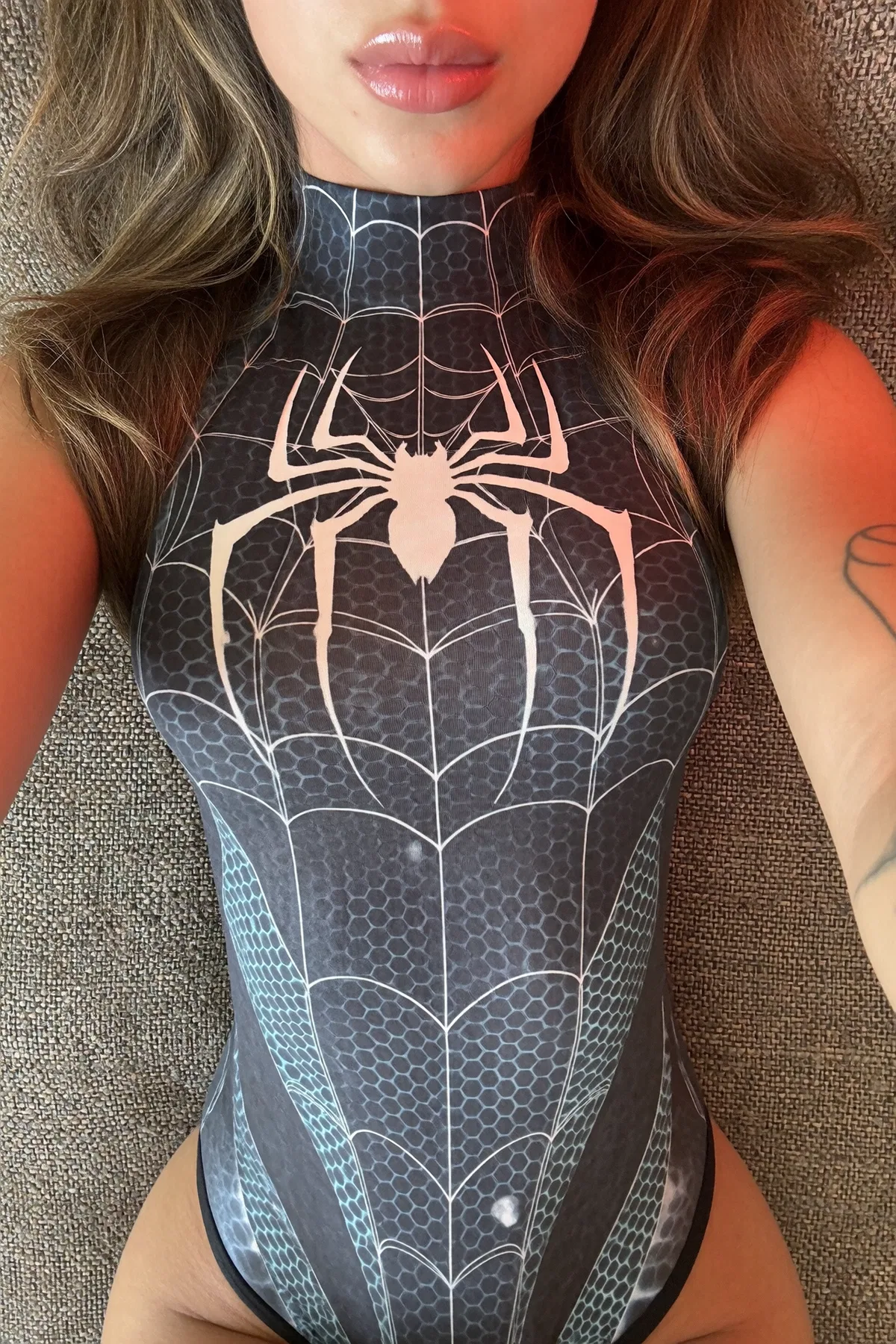 Girl in a dark spiderweb-patterned bodysuit with long wavy brown hair, glossy lips, and an arm tattoo