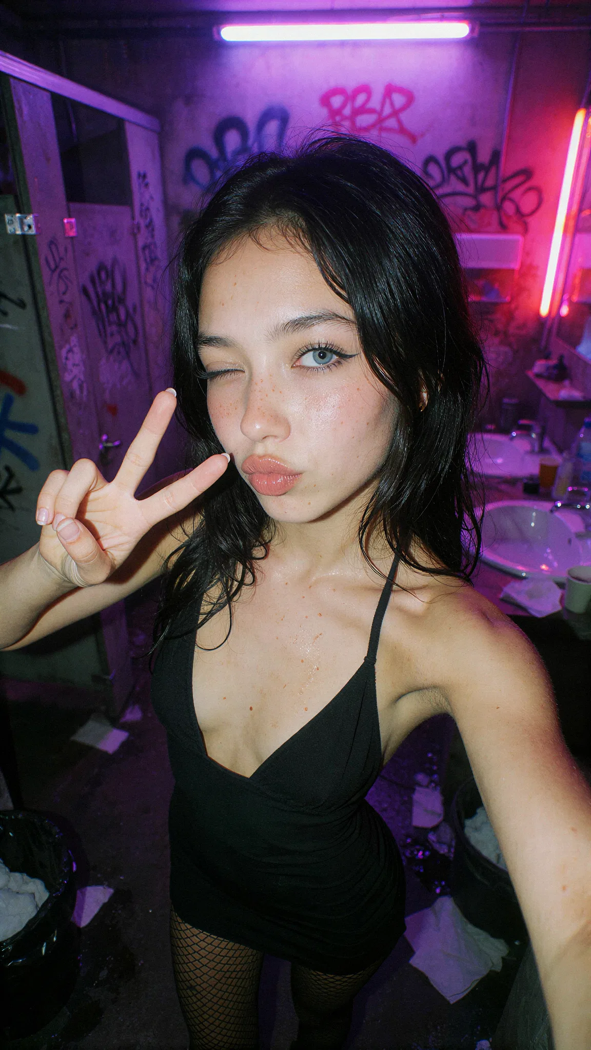 Girl winking and making a peace sign in a black outfit beneath purple and pink neon lights