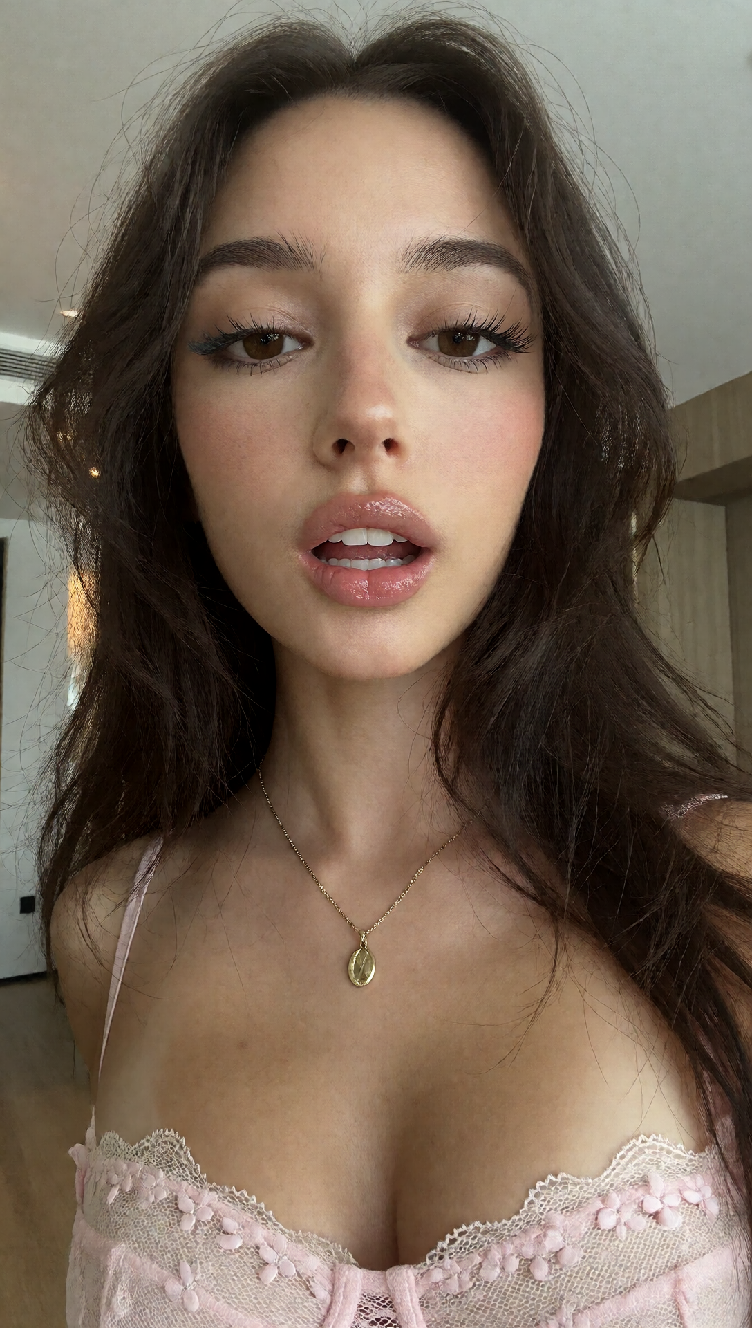 Close-up selfie of a dark-haired girl in a pale pink lace-trimmed camisole and pendant necklace indoors